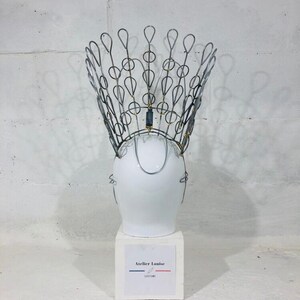 May include: A white ceramic mannequin head wearing a silver wire crown with gold accents. The crown is shaped like a peacock's tail feathers. The mannequin head is on a white pedestal with a label that reads "Atelier Louise Costume".