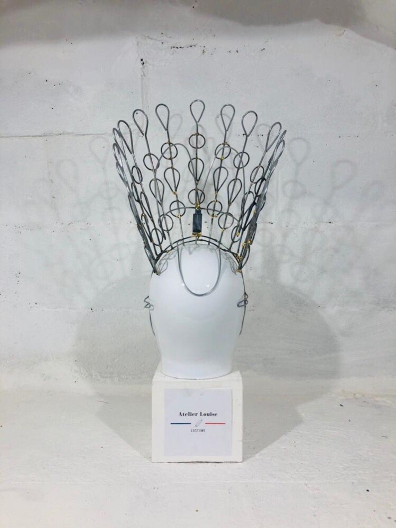 May include: A white ceramic mannequin head wearing a silver wire crown with gold accents. The crown is shaped like a peacock's tail feathers. The mannequin head is on a white pedestal with a label that reads "Atelier Louise Costume".