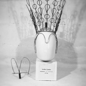 May include: A black and white photo of a wire crown on a white mannequin head. The crown is made of wire circles and loops. The mannequin head is on a white pedestal with a sign that reads "Atelier Louise Costume".