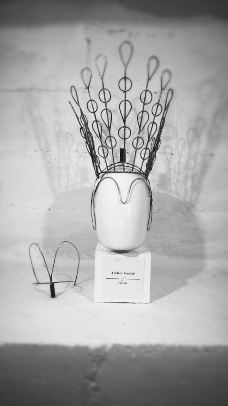 May include: A black and white photo of a wire crown on a white mannequin head. The crown is made of wire circles and loops. The mannequin head is on a white pedestal with a sign that reads "Atelier Louise Costume".