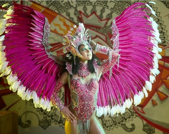 May include: A woman wearing a pink and white feathered costume with a jeweled headpiece. The costume is decorated with rhinestones and sequins. The woman is posing with her arms raised above her head.
