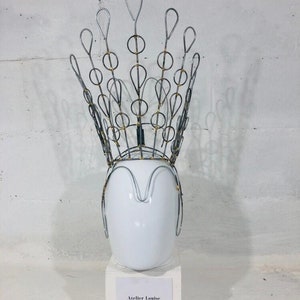 May include: A silver wire crown with gold accents on a white mannequin head. The crown is shaped like a peacock feather with a series of loops and circles. The mannequin head is on a white pedestal with a label that reads "Atelier Louise Costume".