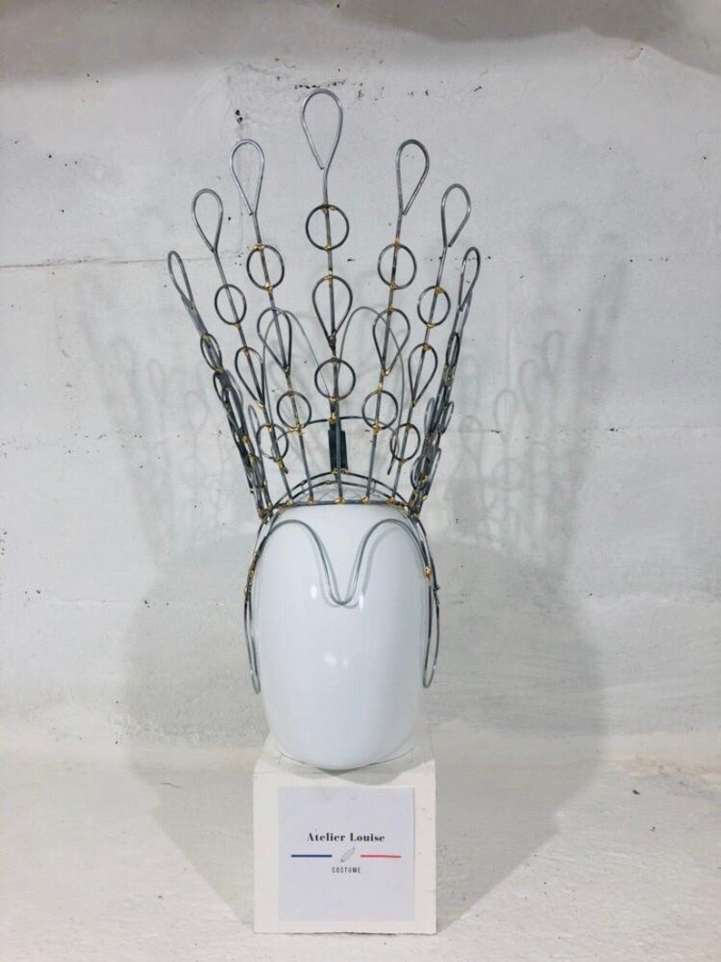May include: A silver wire crown with gold accents on a white mannequin head. The crown is shaped like a peacock feather with a series of loops and circles. The mannequin head is on a white pedestal with a label that reads "Atelier Louise Costume".