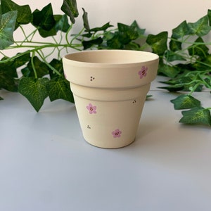 Puede incluir: Una maceta de cerámica de color crema con diseños florales y de puntos rosas. La maceta tiene una forma clásica con un borde ligeramente ensanchado. En el fondo hay vides verdes y frondosas.