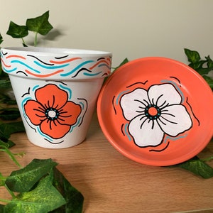 Op de afbeelding: Witte keramische bloempot en bijpassende schotel, beide handbeschilderd met een levendig oranje bloemdesign. De pot heeft een golvend patroon in oranje, turquoise en zwart bij de rand. De schotel is koraalkleurig.