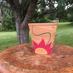 May include: A terracotta-colored flower pot with a hand-painted floral design. The design features a large pink flower with yellow accents, green stems, and orange and yellow dots. The pot is sitting on a wooden table.