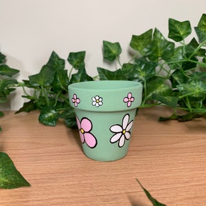 Op de afbeelding: Een kleine, lichtgroene keramische bloempot met handgeschilderde roze en witte madeliefjesontwerpen. De pot heeft een afgeronde vorm en zwarte contouren. De achtergrond heeft groen klimop en een houten oppervlak.