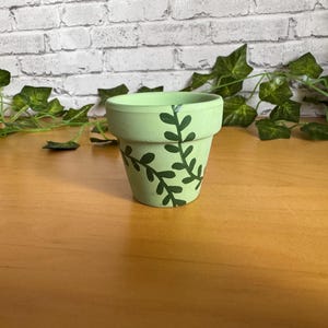 May include: A small, light green painted terracotta flower pot with a dark green leaf design. The pot is cylindrical with a slightly flared rim. The background includes a brick wall and green leafy vines.