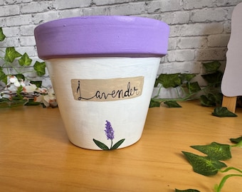 Könnte beinhalten: Ein weiß bemalter Blumentopf mit einem lavendelfarbenen Rand. Der Topf hat ein Etikett mit der Aufschrift "Lavender" und einen gemalten Lavendelzweig. Der Topf steht auf einer Holzoberfläche.