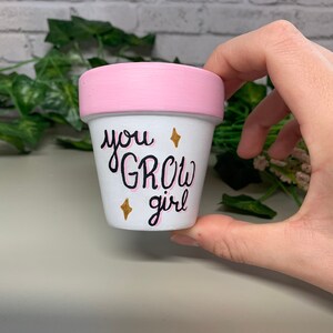 Può includere: Un piccolo vaso di fiori in ceramica bianca con bordo rosa. Il vaso è decorato con le parole "you GROW girl" in scrittura nera con ombreggiatura rosa e dettagli a stella dorata. Ideale per piccole piante o succulente.