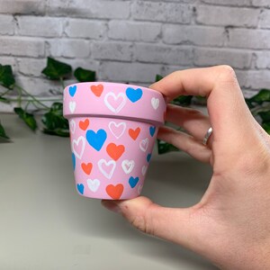 Op de afbeelding: Een kleine, roze beschilderde bloempot versierd met een patroon van witte, oranje en blauwe harten. De pot wordt in een hand gehouden, waarbij het geschilderde ontwerp wordt getoond. Ideaal voor kleine planten of vetplanten.