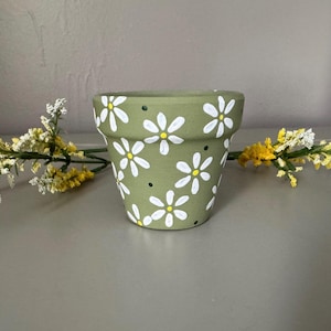 Könnte beinhalten: Kleiner, olivgrüner Keramik-Blumentopf, verziert mit weißen Gänseblümchen und kleinen schwarzen Punkten. Der Topf ist von Zweigen mit weißen und gelben Blüten umgeben, was eine fröhliche, frühlingshafte Ästhetik erzeugt. Ideal für kleine Pflanzen oder Sukkulenten.