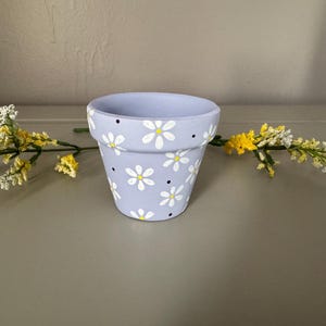 以下が含まれることがあります： 白いデイジーと黒いドットで飾られた、小さな薄紫色の植木鉢。 植木鉢は、黄色と白の花のアクセントが付いたニュートラルな表面に置かれています。