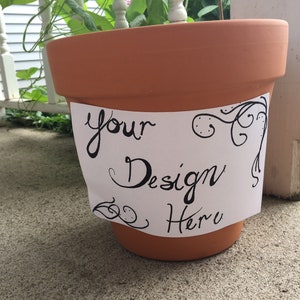 May include: A terracotta-colored flower pot with a white label that reads "your Design Here" in black script. The label has decorative black swirls. The pot is sitting on a concrete surface.