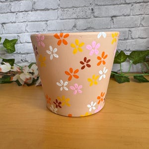 Può includere: Un vaso di fiori in ceramica beige decorato con fiori dipinti a mano in arancione, giallo, rosa, bianco e marrone. Il vaso ha una forma leggermente svasata ed è adatto per uso interno o esterno.