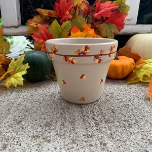 Puede incluir: Una maceta de cerámica beige decorada con un diseño de ramas marrones y hojas de otoño coloridas. La maceta está rodeada de follaje otoñal, calabazas y calabacines, creando una exhibición de temporada. La maceta mide aproximadamente 15 cm de alto.