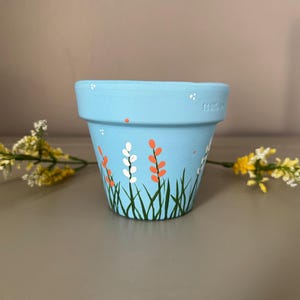 以下が含まれることがあります： 水色のセラミック植木鉢。オレンジ、白、緑の花と草が描かれています。小さな白いドットで装飾され、わずかにフレアしたリムがあります。