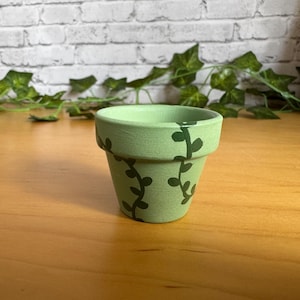 May include: A small, light green ceramic plant pot with dark green vine detailing. The pot is cylindrical with a slightly flared rim. The background features a wooden surface and a white brick wall with green ivy.
