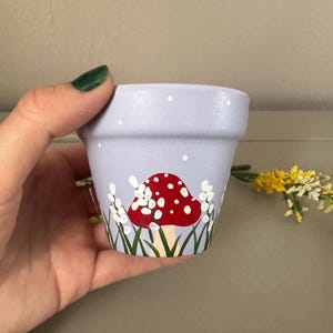 以下が含まれることがあります： 小さな薄紫色のテラコッタ植木鉢。白い水玉模様の赤いキノコ、白い花、緑の草を手描きで描いています。白い点が鉢全体に散らばっています。黄色の花の小枝が右側にあります。