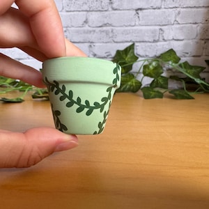 Puede incluir: Una pequeña maceta de terracota pintada de verde claro con un diseño de vid verde oscuro. La maceta se sostiene en una mano, con una superficie de madera y vegetación artificial en el fondo. Ideal para plantas pequeñas.