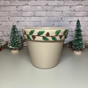 May include: A beige ceramic plant pot with a hand-painted design of green pine needles and brown pine cones around the rim. The pot is surrounded by small Christmas trees and greenery.