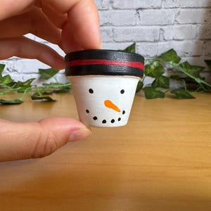 Pode incluir: Um pequeno vaso de cerâmica pintado à mão com tema de boneco de neve. O vaso é branco com uma borda preta e uma faixa vermelha. Apresenta um rosto pintado com pontos pretos para os olhos e boca, e um nariz de cenoura laranja. O vaso é segurado em uma mão.