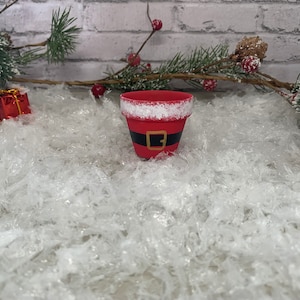 May include: A red painted flower pot decorated to resemble Santa Claus's suit, with a black belt and gold buckle. The rim is trimmed with white paint to mimic snow. The pot is surrounded by decorative holiday elements.