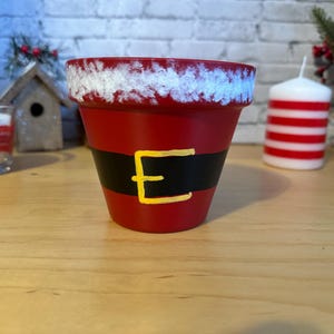 May include: A red terracotta flower pot painted to resemble Santa Claus. The pot has a black belt with a yellow buckle and a white, textured rim. A festive holiday decoration.
