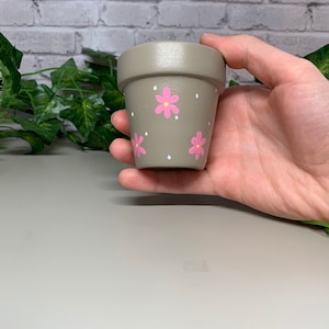 Könnte beinhalten: Ein kleiner, grauer Keramik-Blumentopf mit rosa Blumen und weißen Punkten. Der Topf wird in einer Hand gehalten und eignet sich für kleine Pflanzen oder Sukkulenten. Der Topf hat eine einfache, aber charmante Ästhetik.