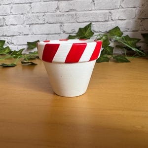 Op de afbeelding: Een kleine, witte keramische bloempot met een rood-witte gestreepte rand. De pot heeft een cilindrische vorm met een licht taps toelopende vorm. De achtergrond omvat een houten oppervlak, een witte bakstenen muur en groen gebladerte.