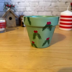 Puede incluir: Maceta de cerámica verde claro pintada a mano, decorada con ramitas de hojas verdes y bayas rojas. La maceta es una decoración festiva, ideal para plantas pequeñas o exhibiciones de temporada.