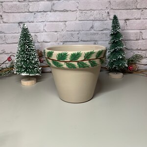 May include: A beige ceramic plant pot with a hand-painted green and brown leaf design around the rim. The pot is surrounded by small decorative Christmas trees and greenery, creating a festive holiday theme.