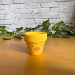 May include: A small, yellow painted terracotta flower pot with a duck face design. The pot has black dot eyes and a pink beak. The pot is sitting on a wooden surface.