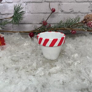May include: A small, white ceramic pot with a red and white striped rim. The pot is set against a backdrop of faux snow, with sprigs of evergreen and red berries. A small, wrapped gift box is also visible.