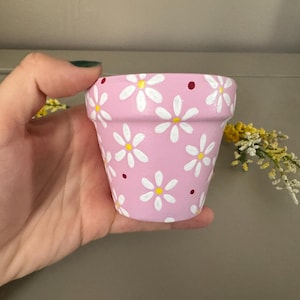 Puede incluir: Una pequeña maceta pintada de color rosa decorada con margaritas blancas y pequeños puntos rojos. La maceta se sostiene en una mano y probablemente se utiliza para plantar pequeñas flores o hierbas. Una adición encantadora a cualquier hogar.