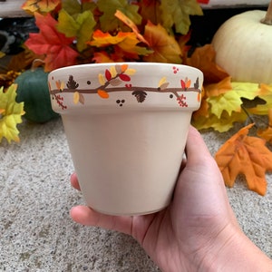 Pode incluir: Um vaso de flores de cerâmica bege com um desenho de outono pintado à mão na borda. O desenho inclui folhas e bagas marrons, laranja e amarelas. O vaso é segurado em uma mão.