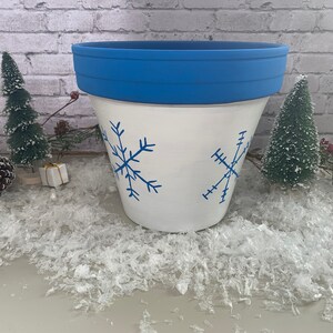 Op de afbeelding: Witte keramische bloempot met een blauwe rand en blauwe sneeuwvlokontwerpen. De pot is versierd met twee blauwe sneeuwvlokken. De pot is omgeven door kunstsneeuw en kleine kerstbomen.