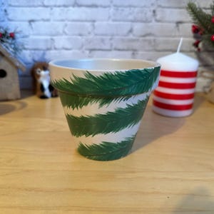 Puede incluir: Maceta de cerámica blanca con un diseño de agujas de pino verdes estilizadas. La maceta es cilíndrica con un borde ligeramente ensanchado. El fondo incluye una pared de ladrillo, una vela y otros elementos decorativos.