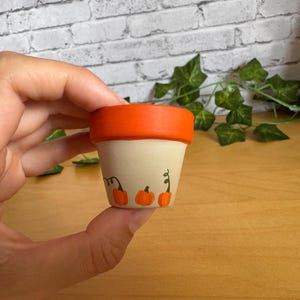May include: A small, hand-painted terracotta flower pot with an orange rim and three orange pumpkins painted on the front. The pot is held in a hand, with a brick wall and greenery in the background.