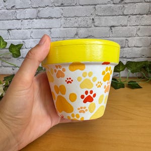 以下が含まれることがあります： 黄色い縁と、オレンジと赤の足跡で飾られた白いボディを持つ、手描きのセラミック植木鉢。 この鉢は、花やハーブを植えるために設計されています。