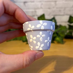 Peut inclure: Un petit pot de fleurs en céramique peint à la main, de couleur lavande clair. Le pot est orné d'un motif répété de fleurs blanches à quatre pétales. Le pot est tenu dans une main, mettant en valeur sa taille et ses détails.