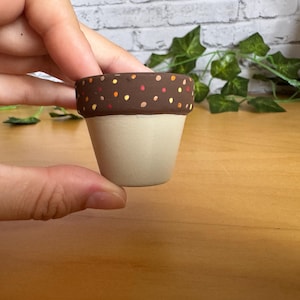 Może przedstawiać: Mały, ręcznie malowany ceramiczny doniczka z jasnobeżową podstawą i ciemnobrązową obwódką. Obwódka jest ozdobiona małymi, kolorowymi kropkami. Doniczka jest trzymana w dłoni, co sugeruje jej rozmiar i zastosowanie do małych roślin lub sukulentów.