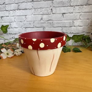 Op de afbeelding: Handbeschilderde keramische bloempot. De pot heeft een crèmekleurige basis met verticale bruine lijnen en een rode rand met witte stippen. Geschikt voor planten, voor binnen of buiten.