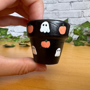 May include: A small, black painted flower pot decorated with white ghost and orange pumpkin designs. The Halloween-themed pot is held in a person's hand and is set against a neutral background.