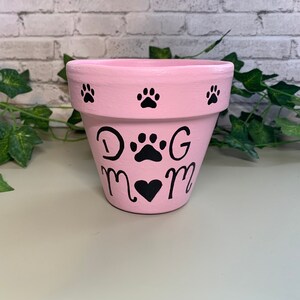 Pode incluir: Um vaso de flores rosa pintado com pegadas pretas e as palavras "DOG MOM" em preto. O vaso tem um símbolo de coração sob as palavras. O vaso está sobre uma superfície clara, com vegetação ao fundo.