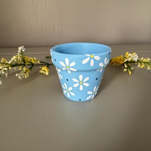 Peut inclure: Un pot de fleurs en céramique bleu clair décoré de marguerites blanches et de petits points bleus. Le pot est cylindrique avec un bord légèrement évasé. Des fleurs artificielles à fleurs jaunes et blanches sont placées près du pot.