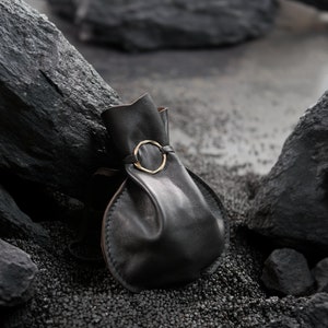 Puede incluir: Una bolsa de cuero negro con cierre de cordón, asegurada por un anillo dorado. La bolsa se encuentra sobre un fondo de rocas oscuras y pequeños guijarros negros, creando una estética minimalista.