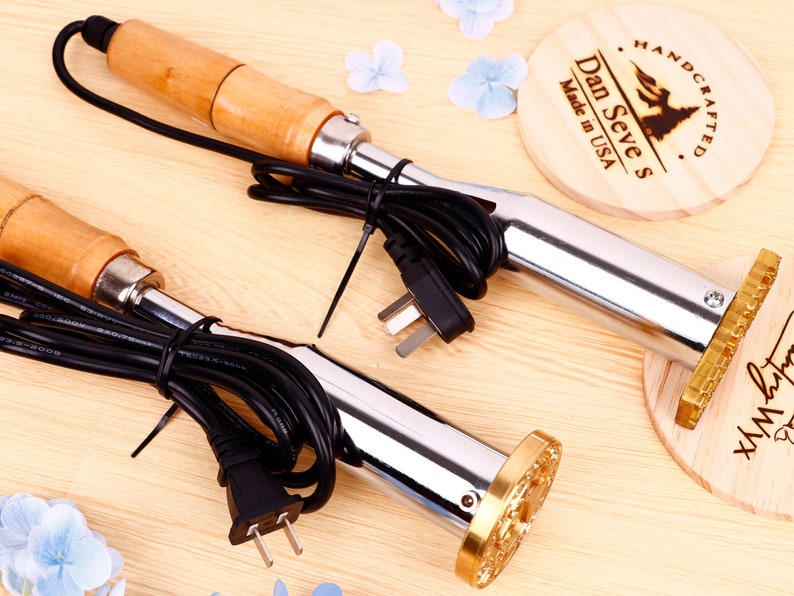 May include: Two electric wood burning tools with wooden handles and black cords. Each tool has a metal tip with a brass-colored branding head. The branding heads are attached to wooden plaques with the text "Dan Seve's Made in USA" and "Handcrafted" on one plaque and a cursive signature on the other.
