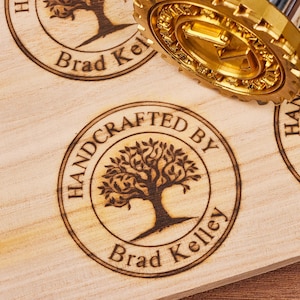 Puede incluir: Primer plano de una superficie de madera con un diseño de sello circular hecho a mano. El sello presenta un árbol estilizado con el texto "HANDCRAFTED BY Brad Kelley" alrededor del perímetro.
