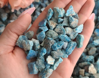 May include: A hand holding a pile of small, blue-green, rough-textured stones. The stones are scattered on a surface covered in similar stones.
